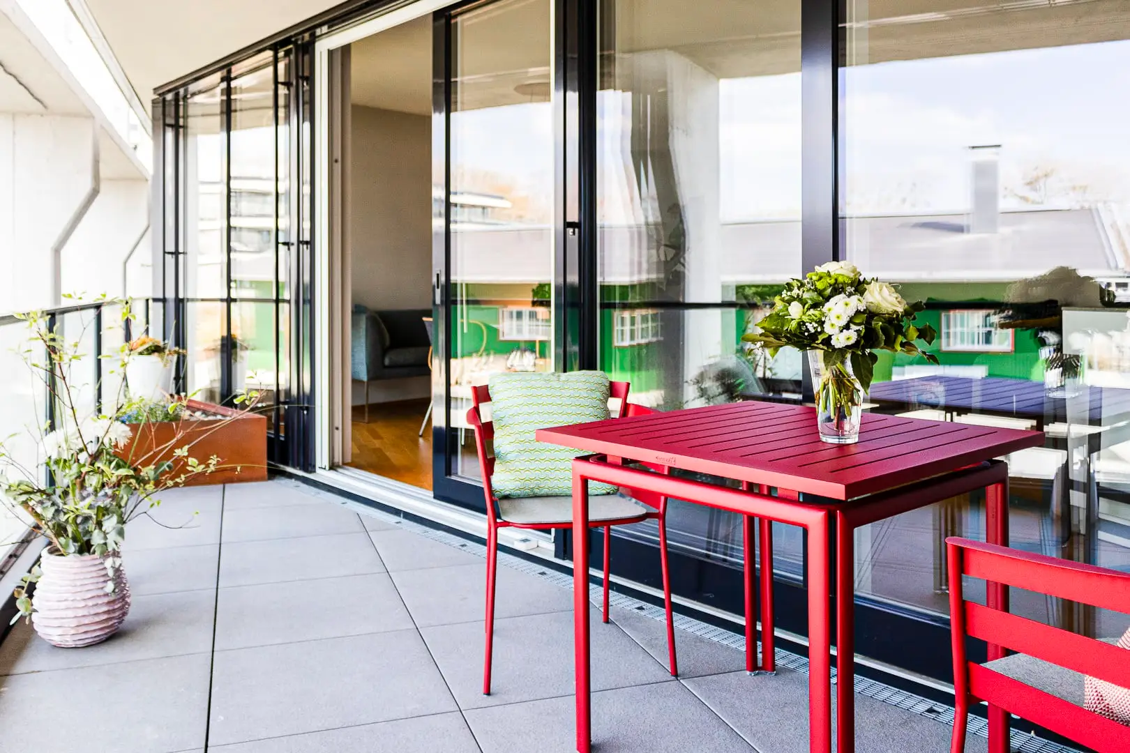 Balkon mit roten Gartenmöbeln. Links sind Pflanzen in Töpfen, und im Hintergrund ist eine große Glas-Schiebetür zu sehen, die den Balkon mit dem Innenraum verbindet.