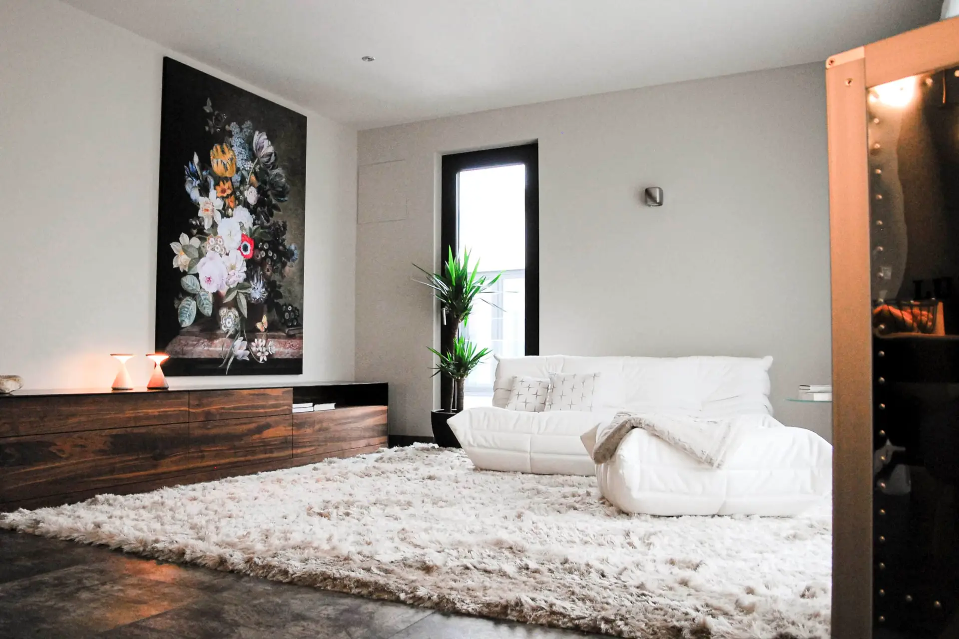 Ein modernes Wohnzimmer mit einem weißen Sofa, einem großen flauschigen Teppich und einer dunklen Holzkommode. An der Wand hängt ein großes Blumenbild. Eine grüne Pflanze steht neben einem Fenster, das Licht in den Raum bringt.