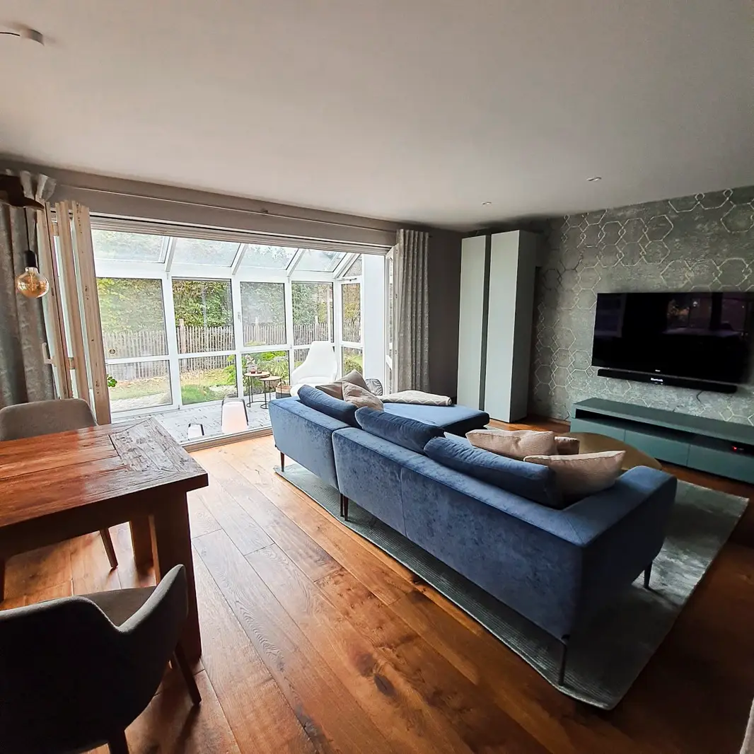 Ein helles Wohnzimmer mit einem blauen Sofa, einem Holztisch und einem großen Fernseher an der Wand. Der Raum öffnet sich zu einem Wintergarten mit Blick auf den Garten.