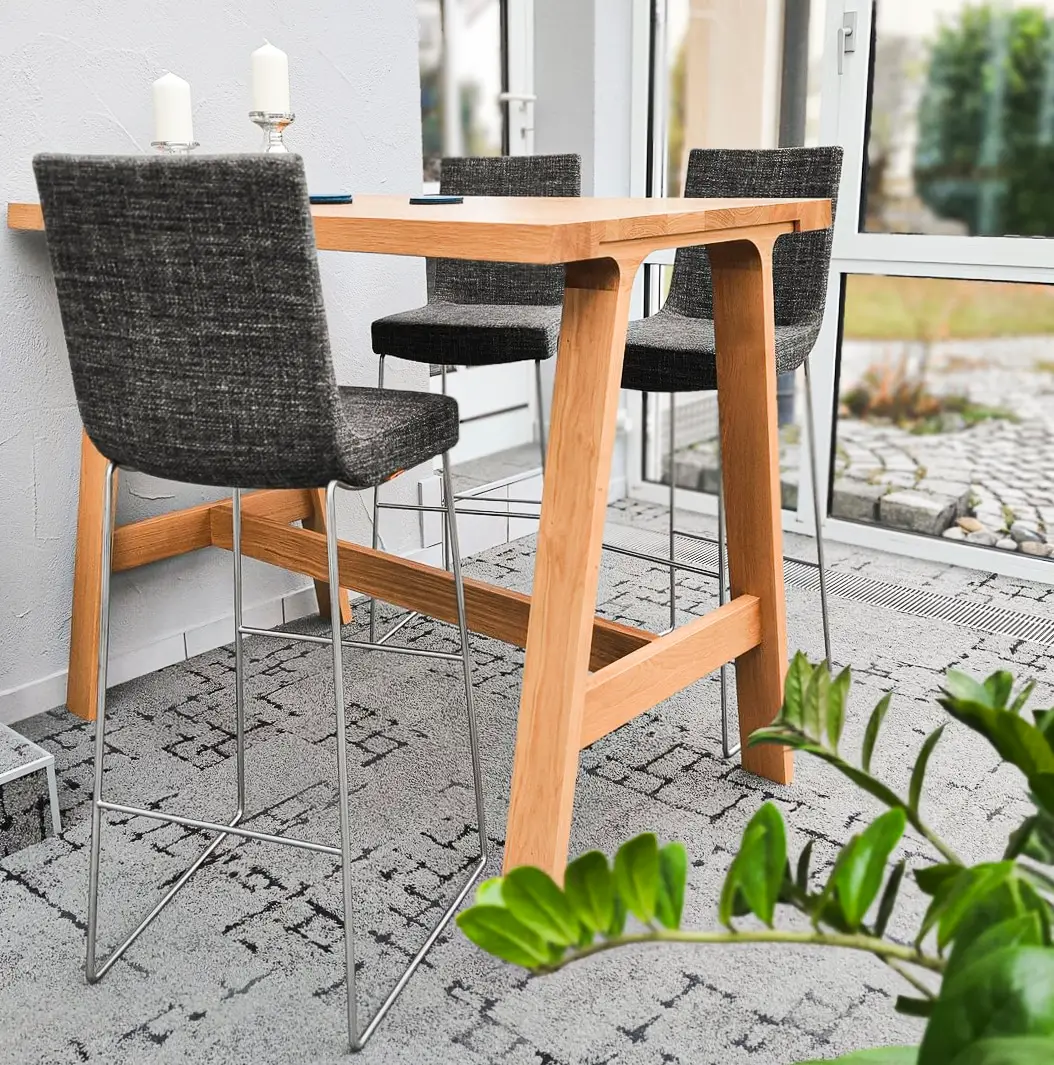 Ein Wintergarten mit einem hohen Holztisch und drei grauen Barhockern. Auf dem Tisch stehen zwei Kerzen, und große Fenster bieten einen Blick nach draußen. Eine Pflanze steht im Vordergrund.