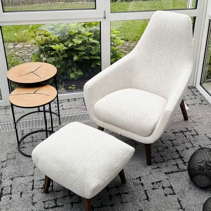Ein heller Sitzbereich mit einem weißen Sessel, Fußhocker, zwei kleinen Holztischen und drei dekorativen Körben. Große Fenster bieten Blick auf den Garten.