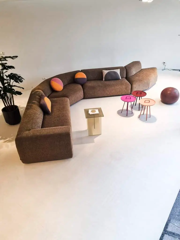 Ein modernes Wohnzimmer mit einem braunen Sofa, bunten Kissen und kleinen, farbigen Beistelltischen. Links steht eine Pflanze in einem Topf.
