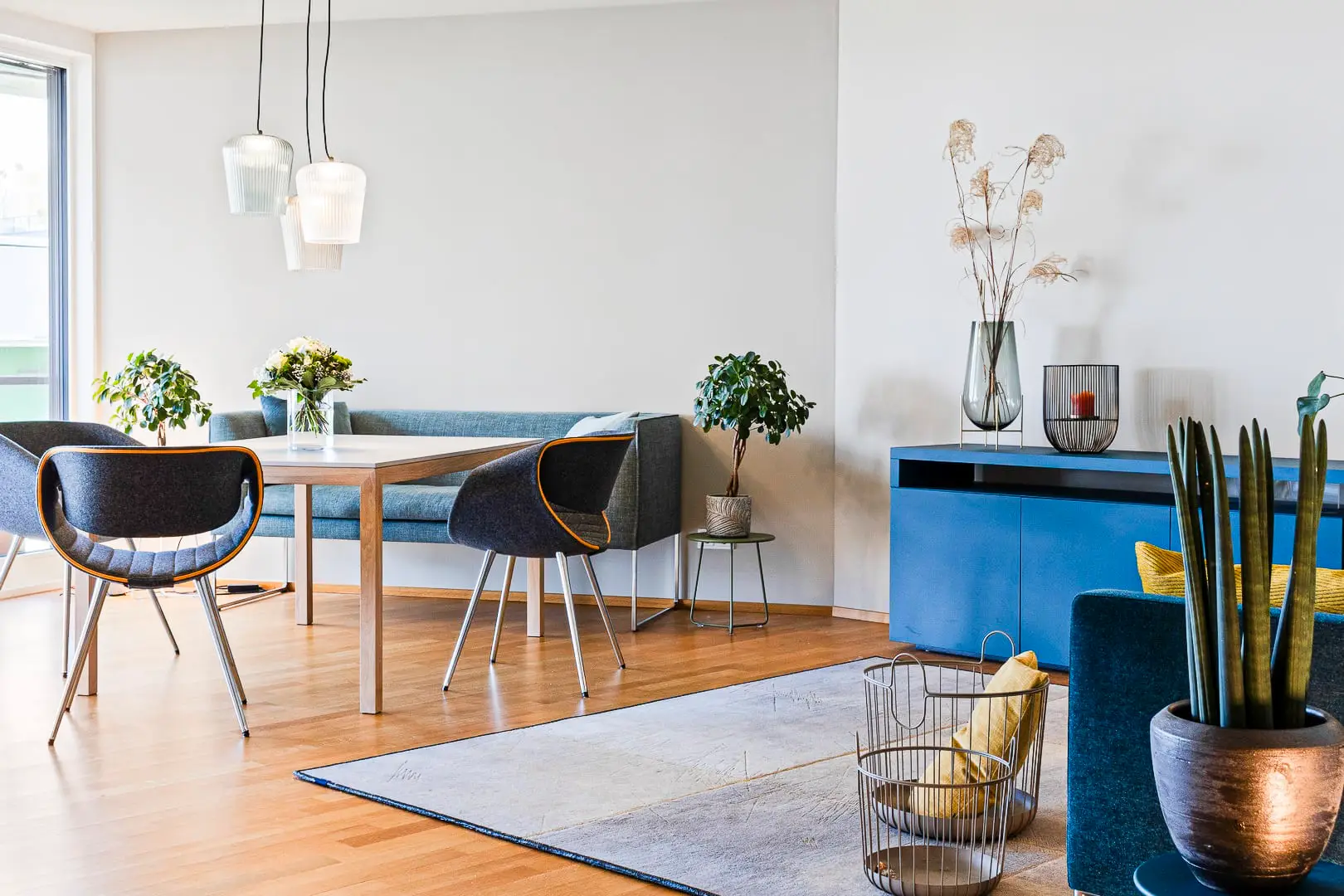 Ein moderner Essbereich mit einem Holztisch, vier blauen Stühlen und hängenden Lampen. Eine Eckbank und Pflanzen verleihen Gemütlichkeit. Ein blaues Sideboard mit Dekorationen und ein großer Teppich runden den Raum ab.
