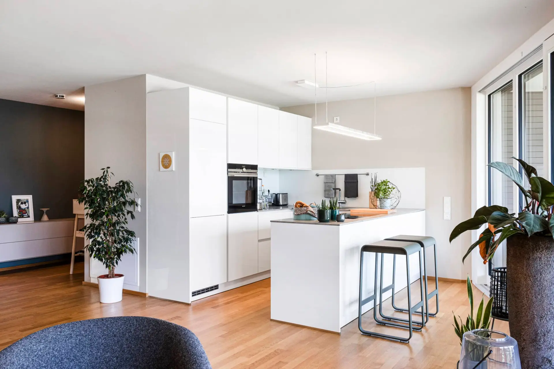 Eine moderne, helle Küche mit weißen Schränken, einer Kücheninsel und zwei Barhockern. Pflanzen und Dekorationsgegenstände sorgen für eine gemütliche Atmosphäre.