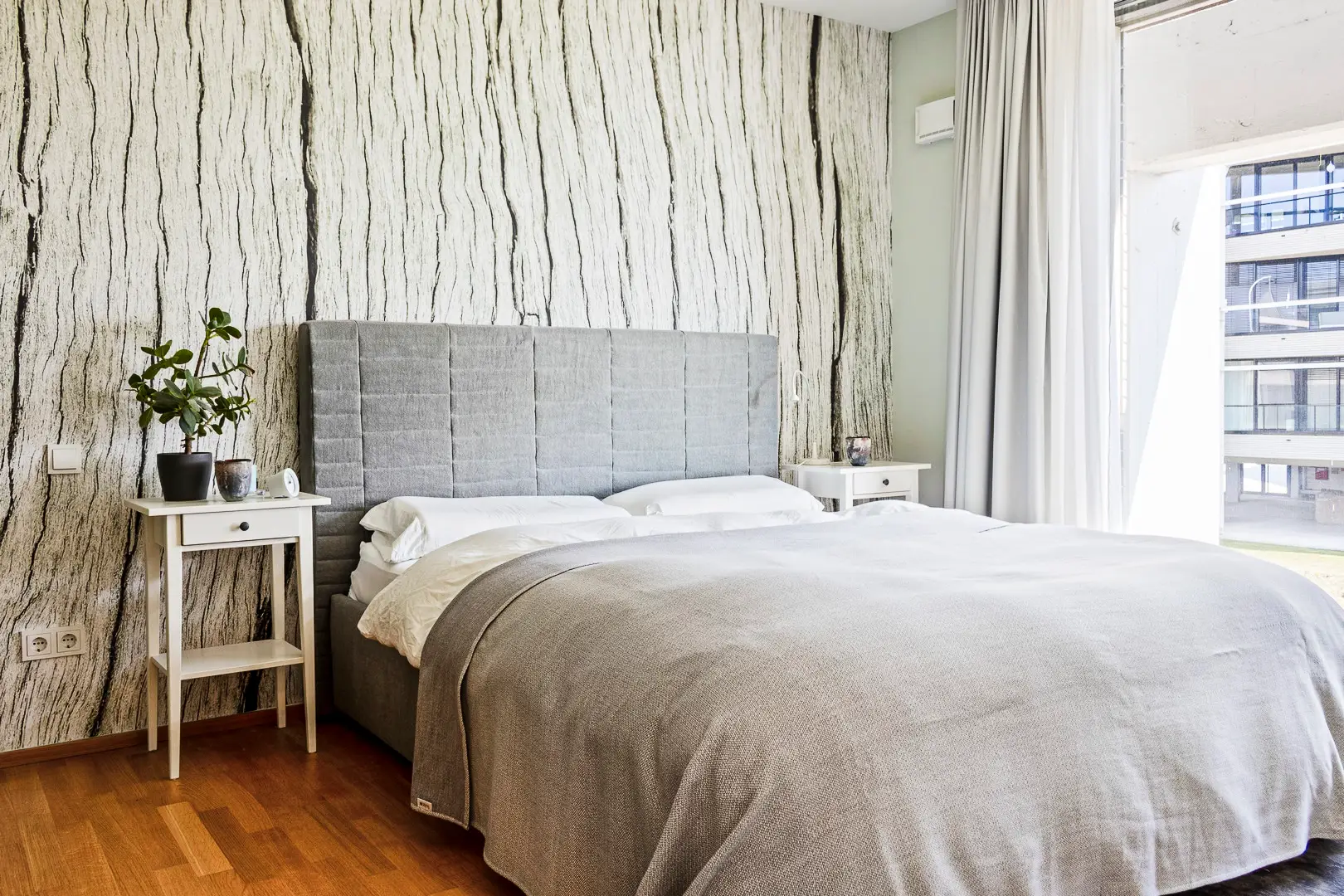Ein helles Schlafzimmer mit einem grauen gepolsterten Bett, weißen Nachttischen und einer Wand mit einem baumrindenartigen Muster. Große Fenster lassen viel Tageslicht herein.