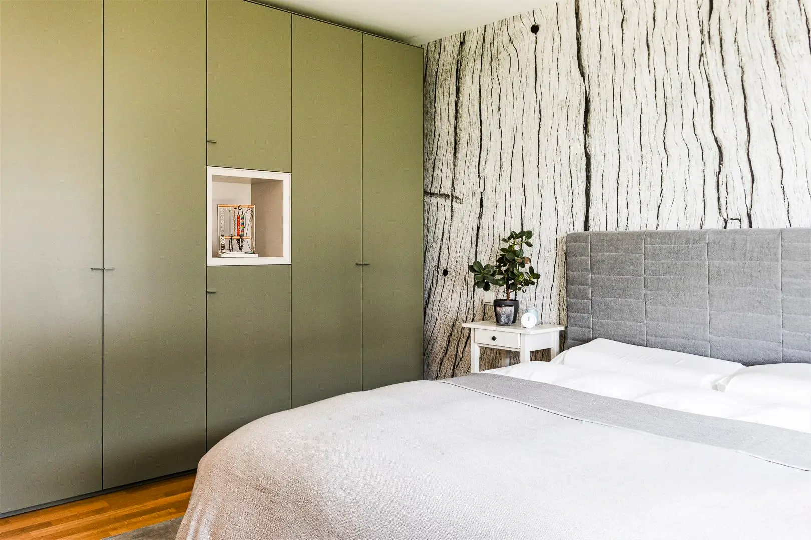 Ein helles Schlafzimmer mit einem grauen gepolsterten Bett, einer baumrindenartigen Akzentwand und olivgrünen Einbauschränken. Ein Nachttisch mit einer Pflanze steht neben dem Bett.