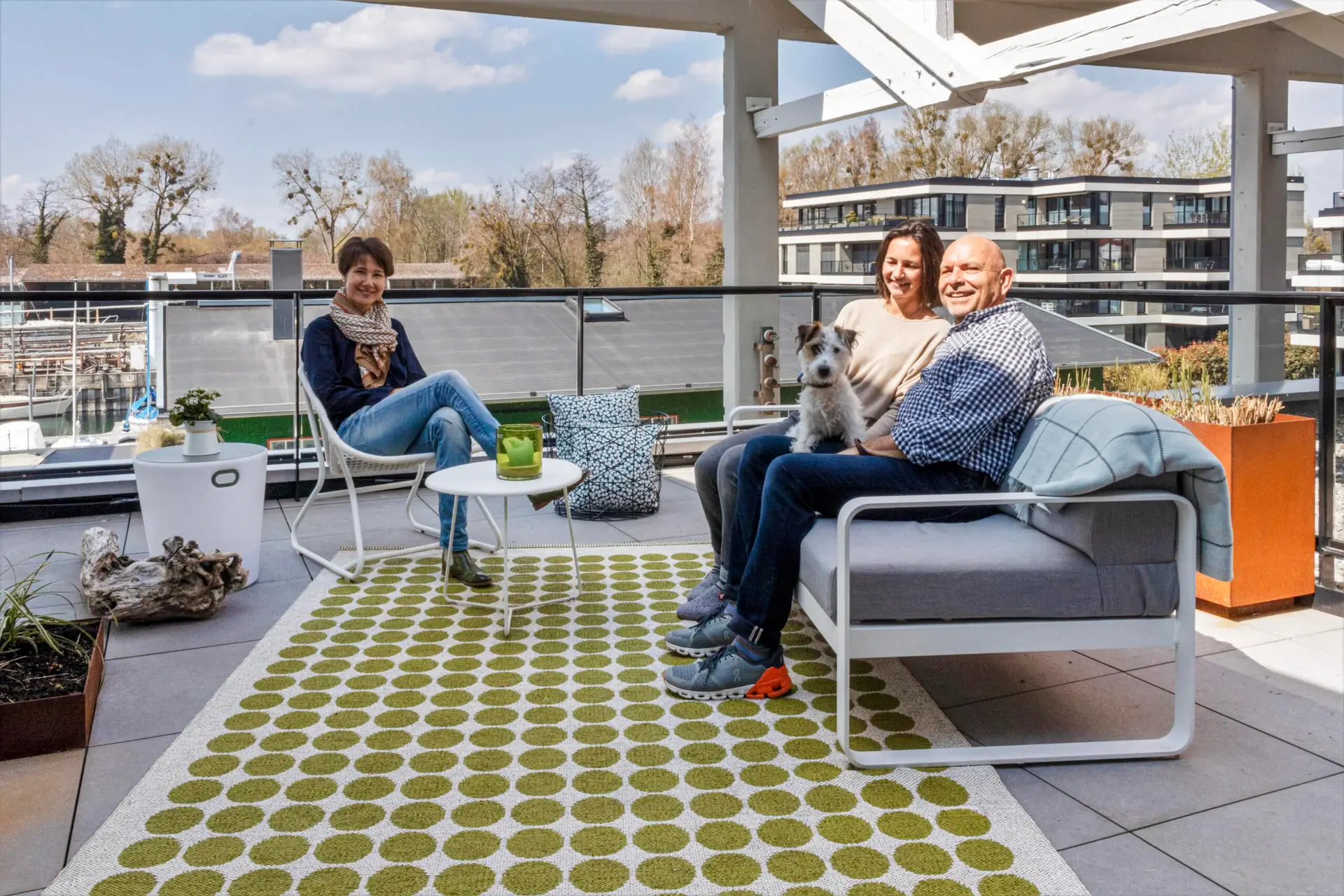 Frau Hafen und ein Pärchen mit einem Hund auf dem Schoß sitzen auf einem Balkon mit viel Dekoration. Sie sitzen auf modernen Möbeln, die auf einem weißen Teppich mit grünen Punkten stehen.