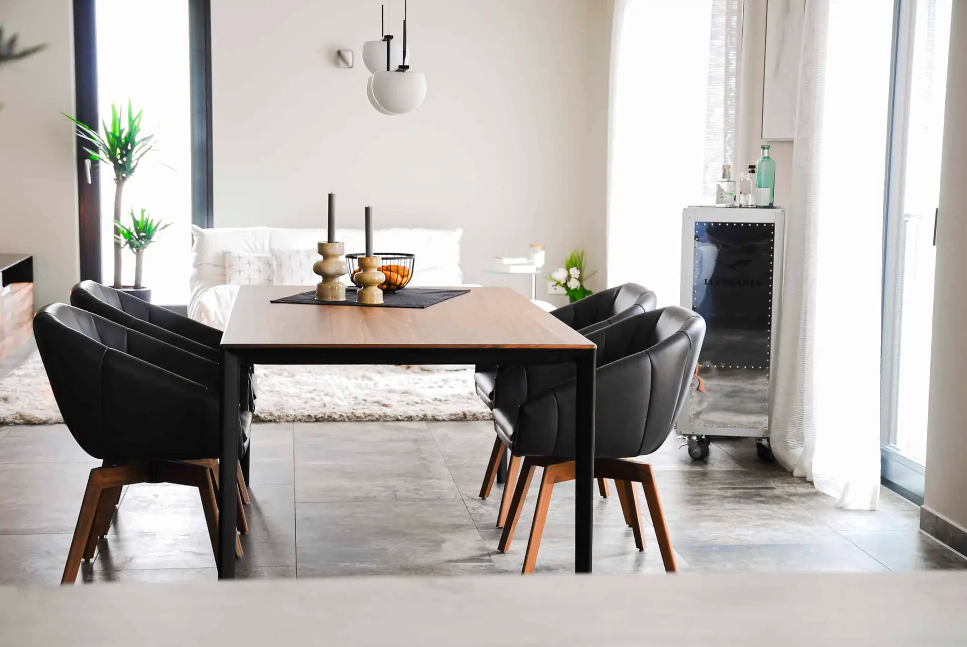 Ein modernes Esszimmer mit einem Holztisch und vier schwarzen Stühlen. Auf dem Tisch stehen Dekorationsobjekte und Kerzen. Im Hintergrund sind ein helles Sofa, eine Zimmerpflanze und große Fenster zu sehen.