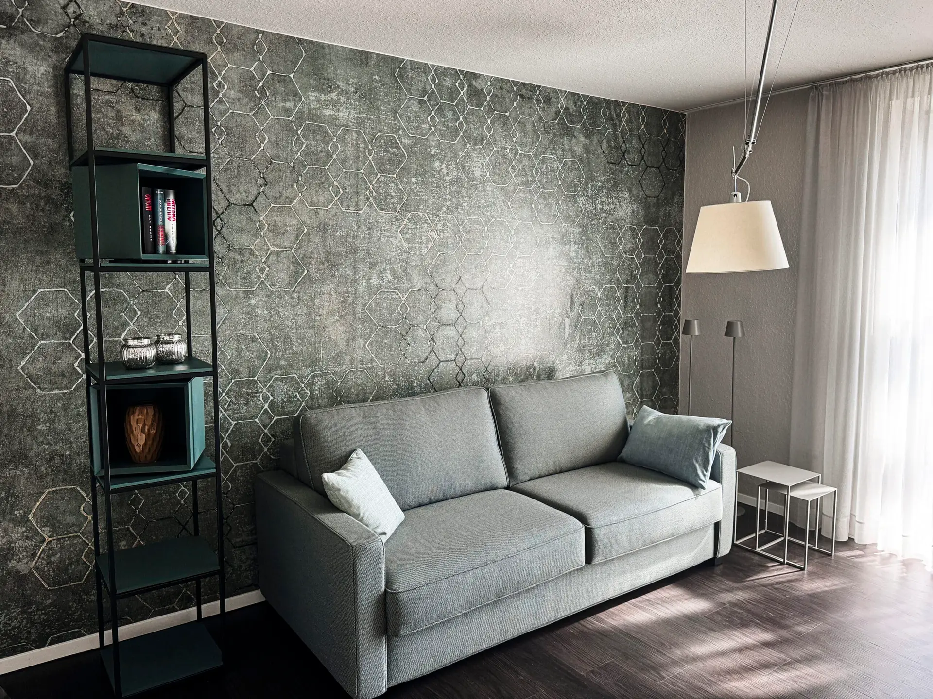 Ein modernes Wohnzimmer mit einem grauen Sofa, einer Wand mit hexagonalem Muster und einem schmalen Regal. Eine große Hängelampe und zwei Beistelltische ergänzen die Einrichtung.
