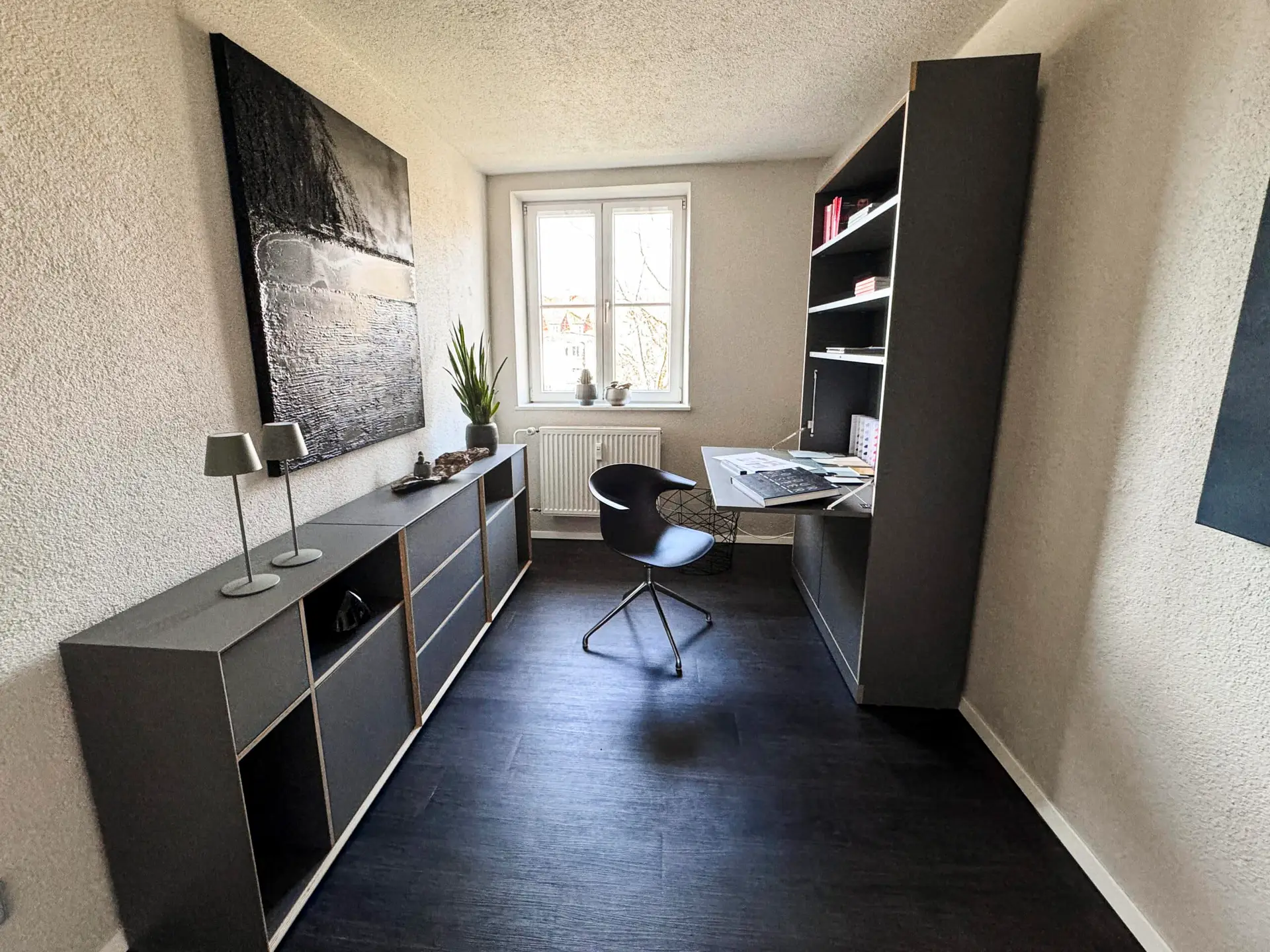 Ein modernes, helles Arbeitszimmer mit einem Schreibtisch, einem schwarzen Stuhl und einem Regal. An der Wand hängt ein großes abstraktes Kunstwerk. Ein Fenster lässt Tageslicht herein.