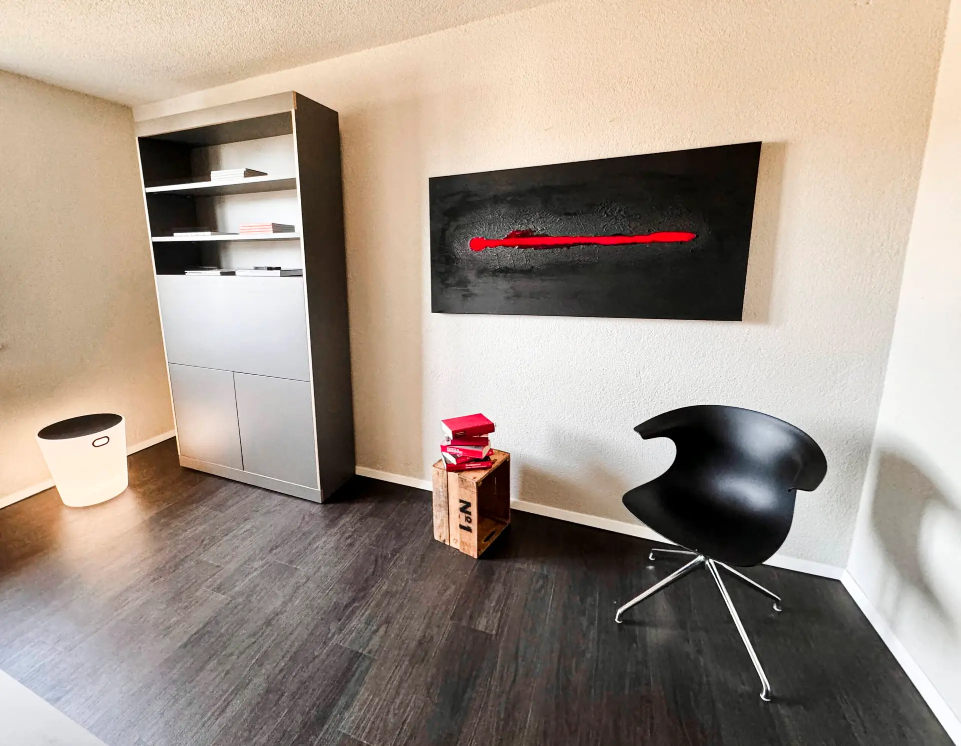 Ein minimalistisches Arbeitszimmer mit einem schwarzen Stuhl, einem hohen Regal und einer modernen Lampe. An der Wand hängt ein Kunstwerk mit einem roten Akzent. Ein Holzkasten dient zur Ablage.