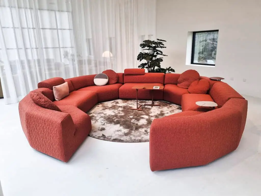 Ein modernes Wohnzimmer mit einem großen, roten, halbkreisförmigen Sofa. In der Mitte liegt ein runder Teppich, darauf stehen kleine Beistelltische. Im Hintergrund sind weiße Vorhänge und eine Pflanze zu sehen.