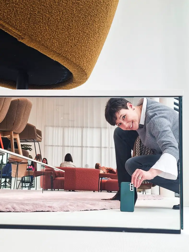 Frau Hafen macht ein Foto von sich selbst in einem Spiegelsockel, während sie in einem modernen, gemütlichen Raum mit bequemen Sesseln und Teppichen ist. Im Hintergrund sitzen mehrere Menschen auf roten Sofas.