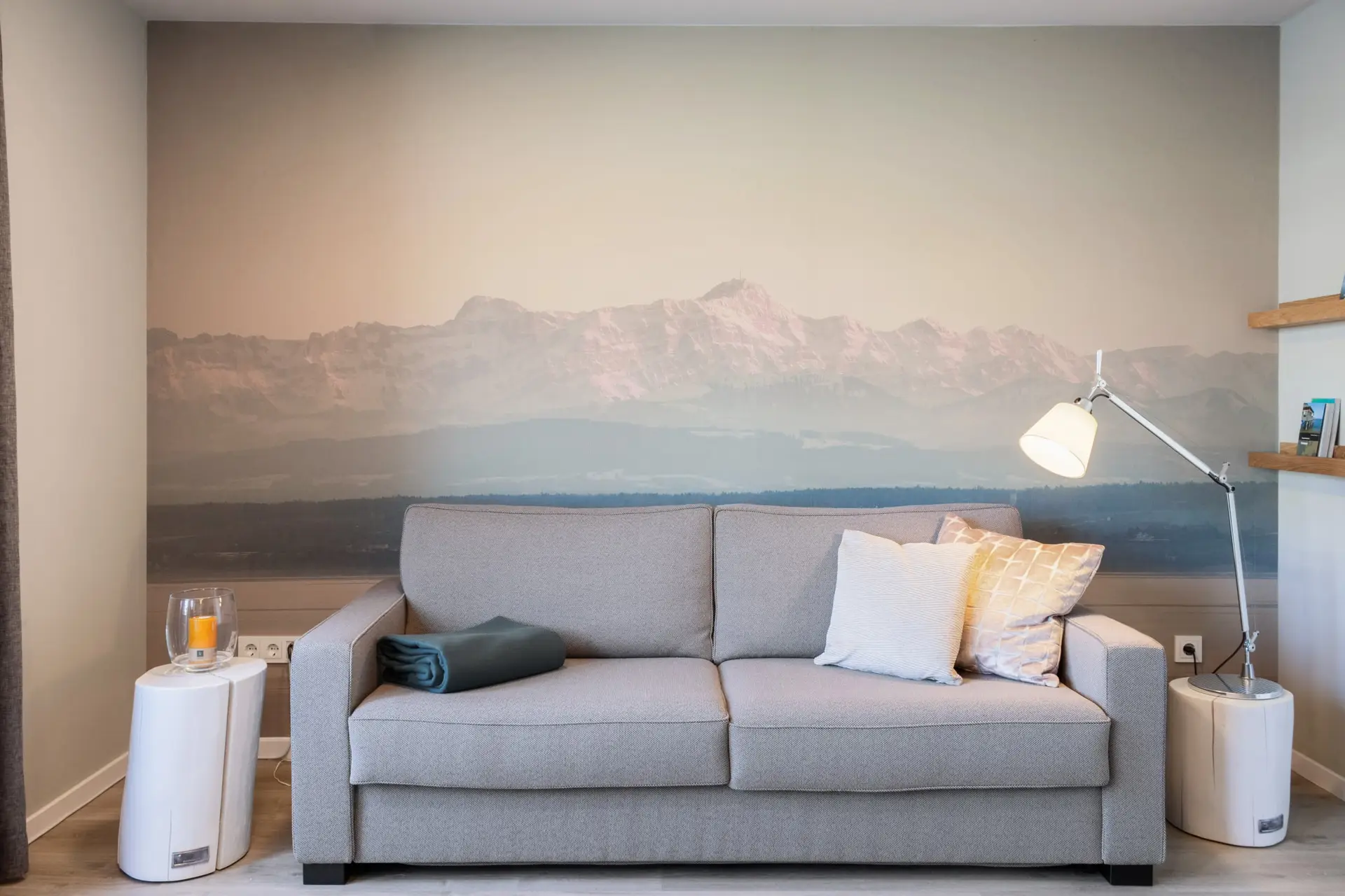 Ein graues Sofa vor einer Wand mit Bergmotiv. Auf beiden Seiten stehen weiße Beistelltische mit einer Kerze und einer Lampe.