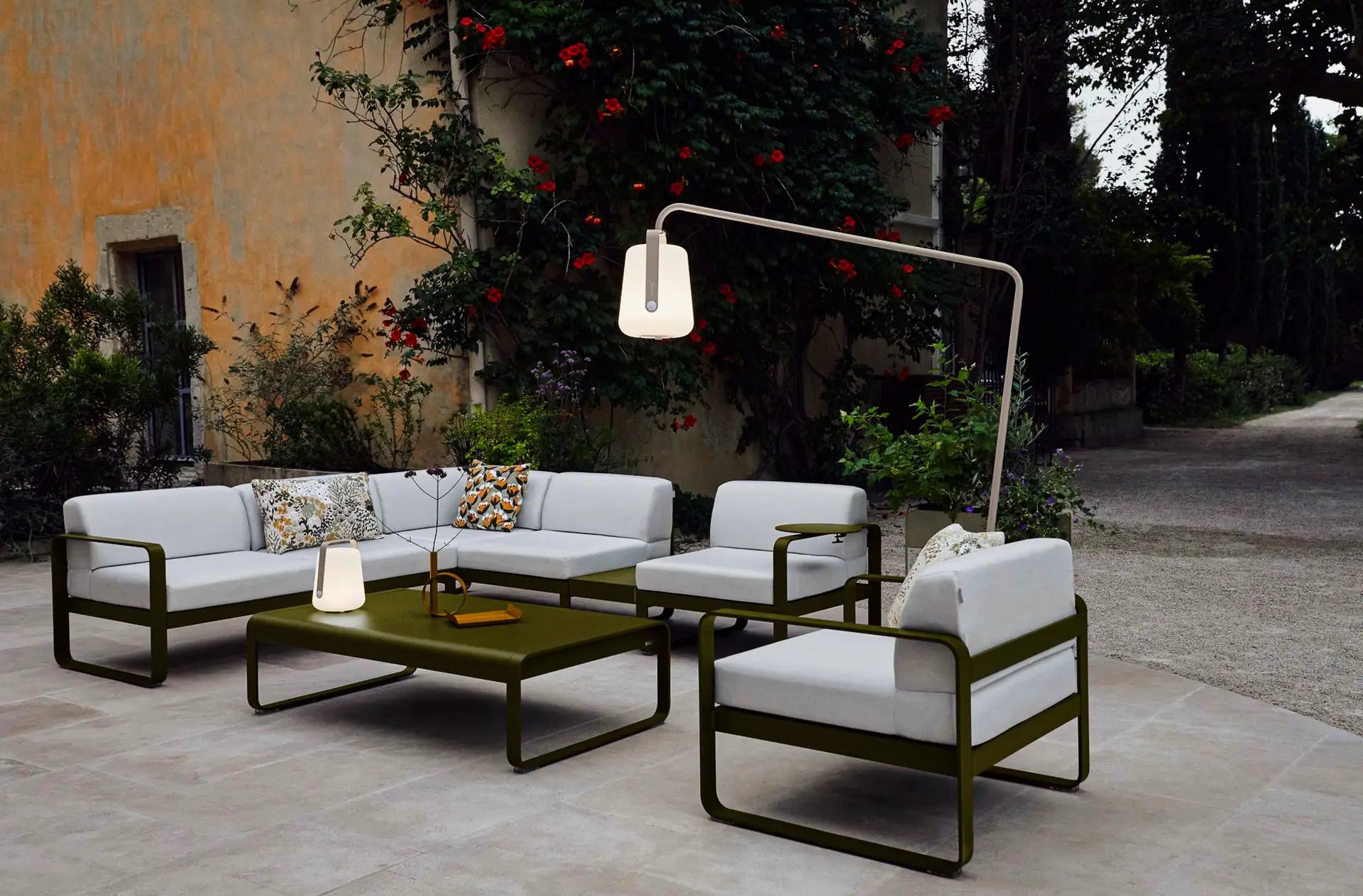 Eine Terrasse mit weiß gepolsterten Sitzmöbeln und olivgrünen Metallrahmen. Ein großer Couchtisch und eine moderne Stehlampe stehen in der Mitte. Im Hintergrund sieht man Pflanzen an einer gelben Hauswand und Bäume dahinter.