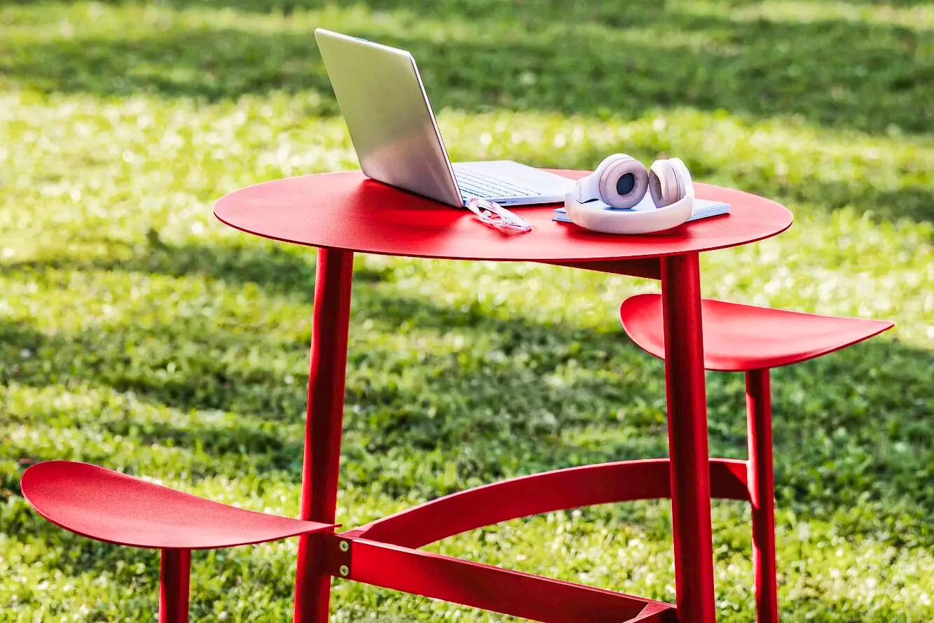 Ein roter Tisch mit zwei integrierten Sitzen steht auf einer grünen Wiese. Auf dem Tisch liegen ein Laptop und Kopfhörer.