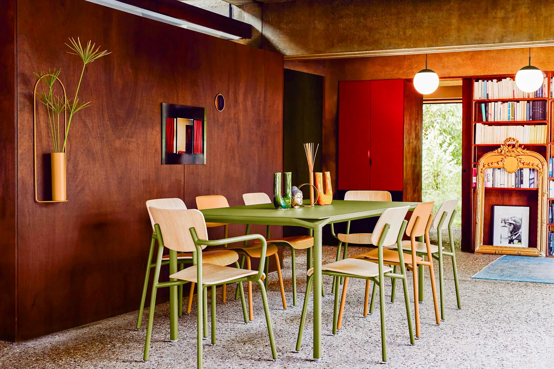 Ein Essbereich mit einem grünen Tisch und Holzstühlen. Die Wand ist aus dunklem Holz und an ihr hängt eine Vase mit Pflanzen, und der Raum ist mit alt-modernen Dekorationen und Büchern ausgestattet.