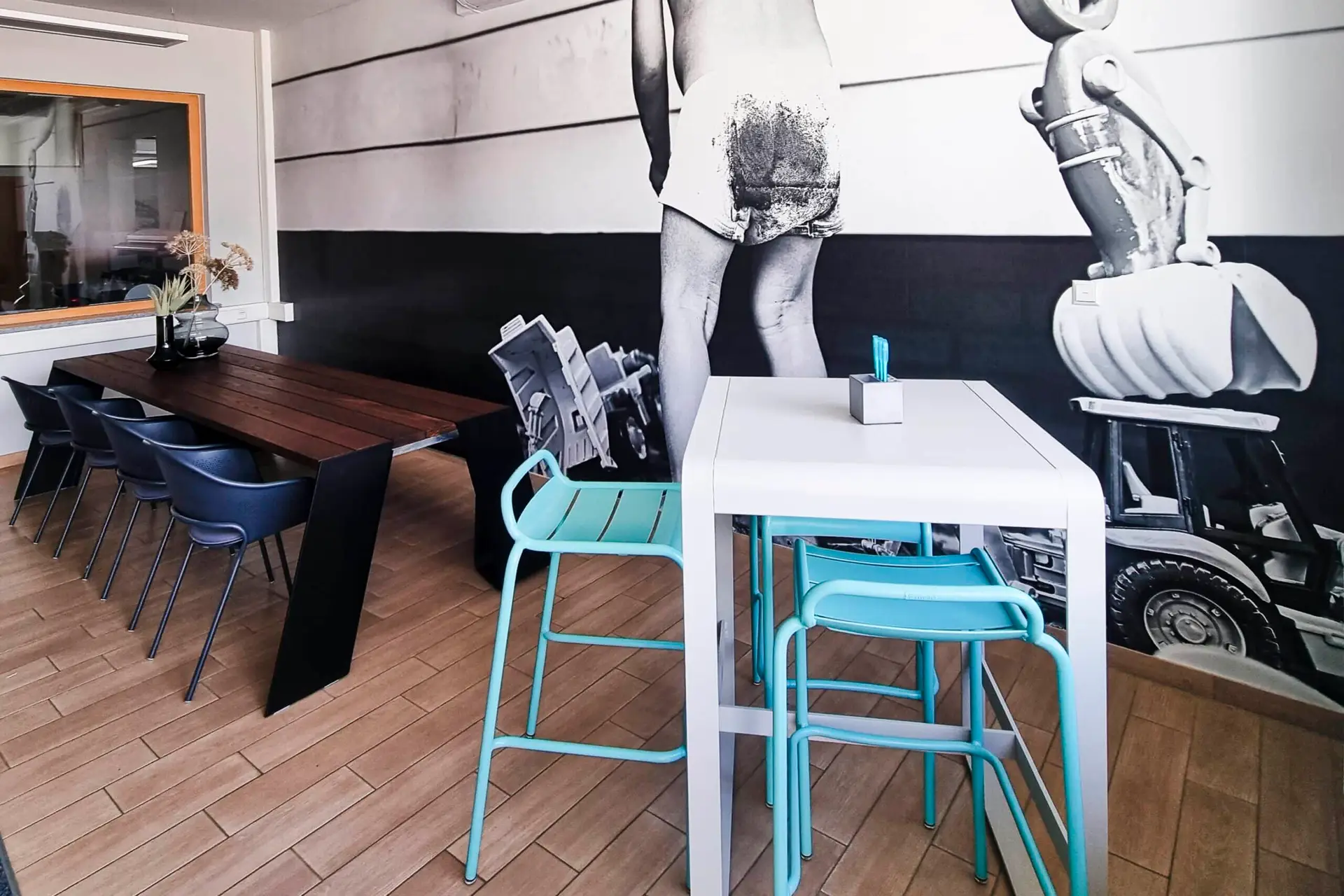 Ein moderner Raum mit einem langen Holztisch und schwarzen Stühlen, daneben ein hoher weißer Tisch mit zwei hellblauen Hockern. An der Wand ist ein großes Schwarz-Weiß-Foto eines Kindes mit einem Spielzeugtraktor.