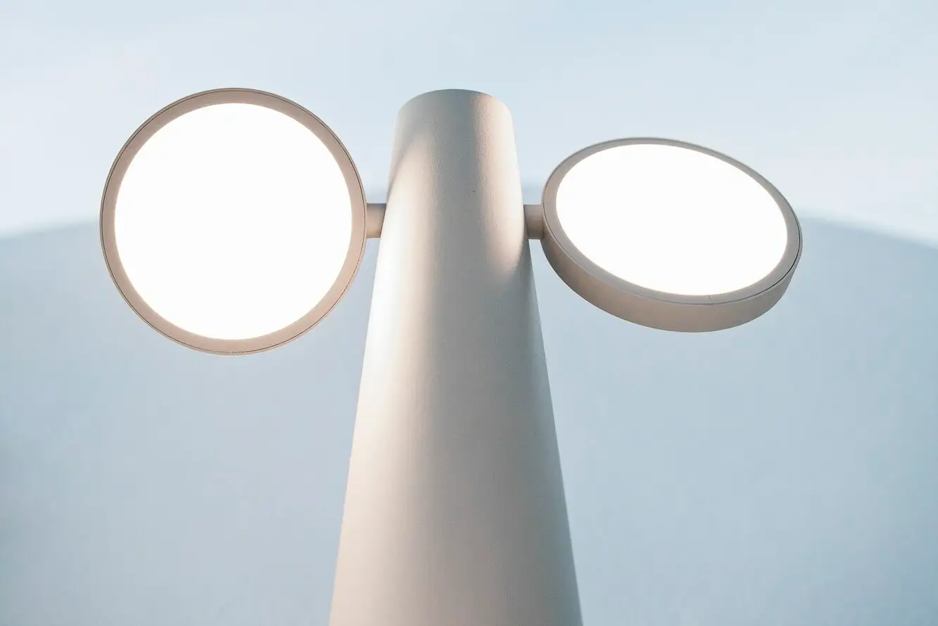 Eine moderne, minimalistische weiße Lampe mit zwei runden, leuchtenden Scheiben auf einem kegelförmigen Sockel vor einem blauen Hintergrund.