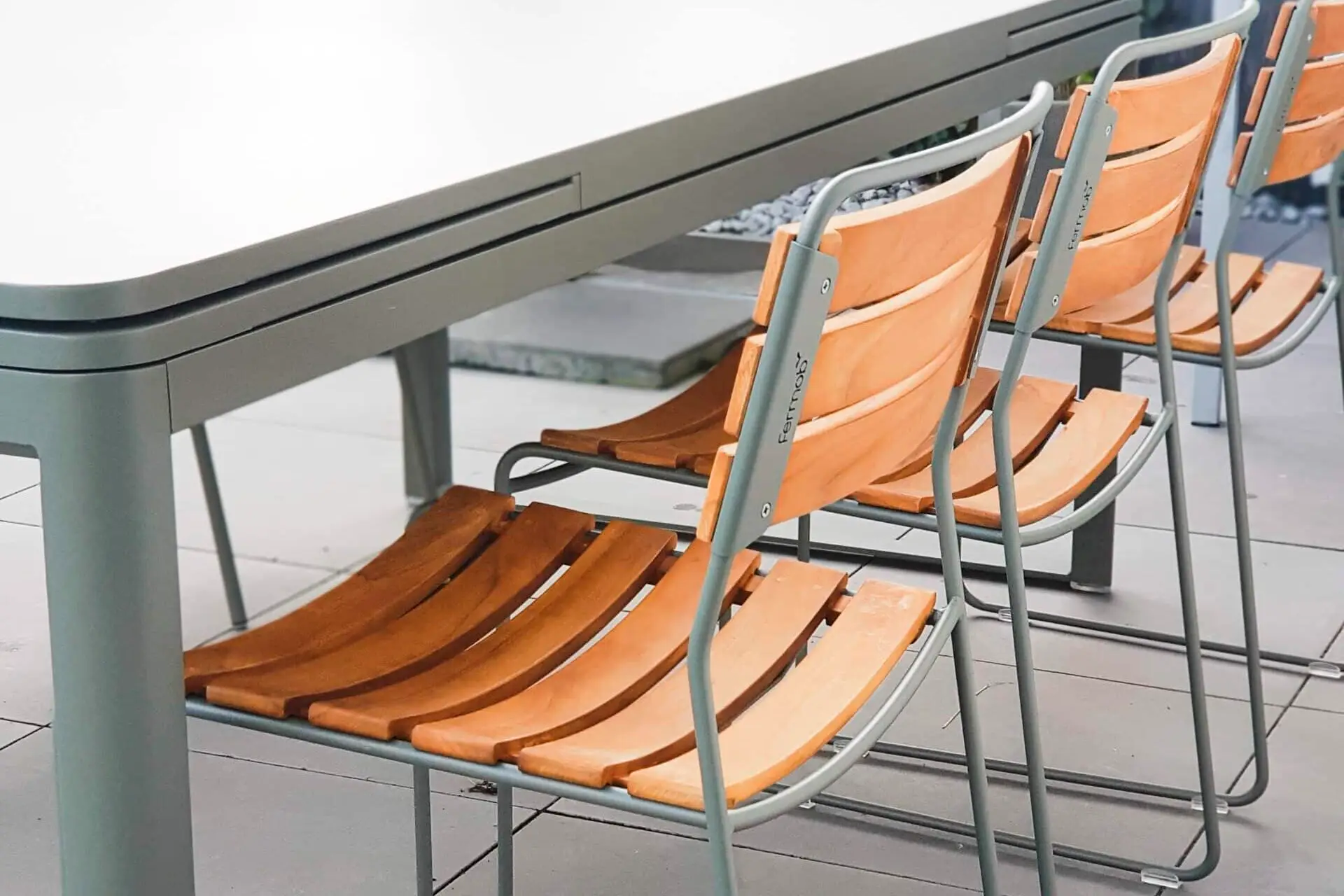 Drei Holzstühle mit grauen Metallrahmen stehen an einem grauen Tisch im Freien, umgeben von grüner Vegetation.