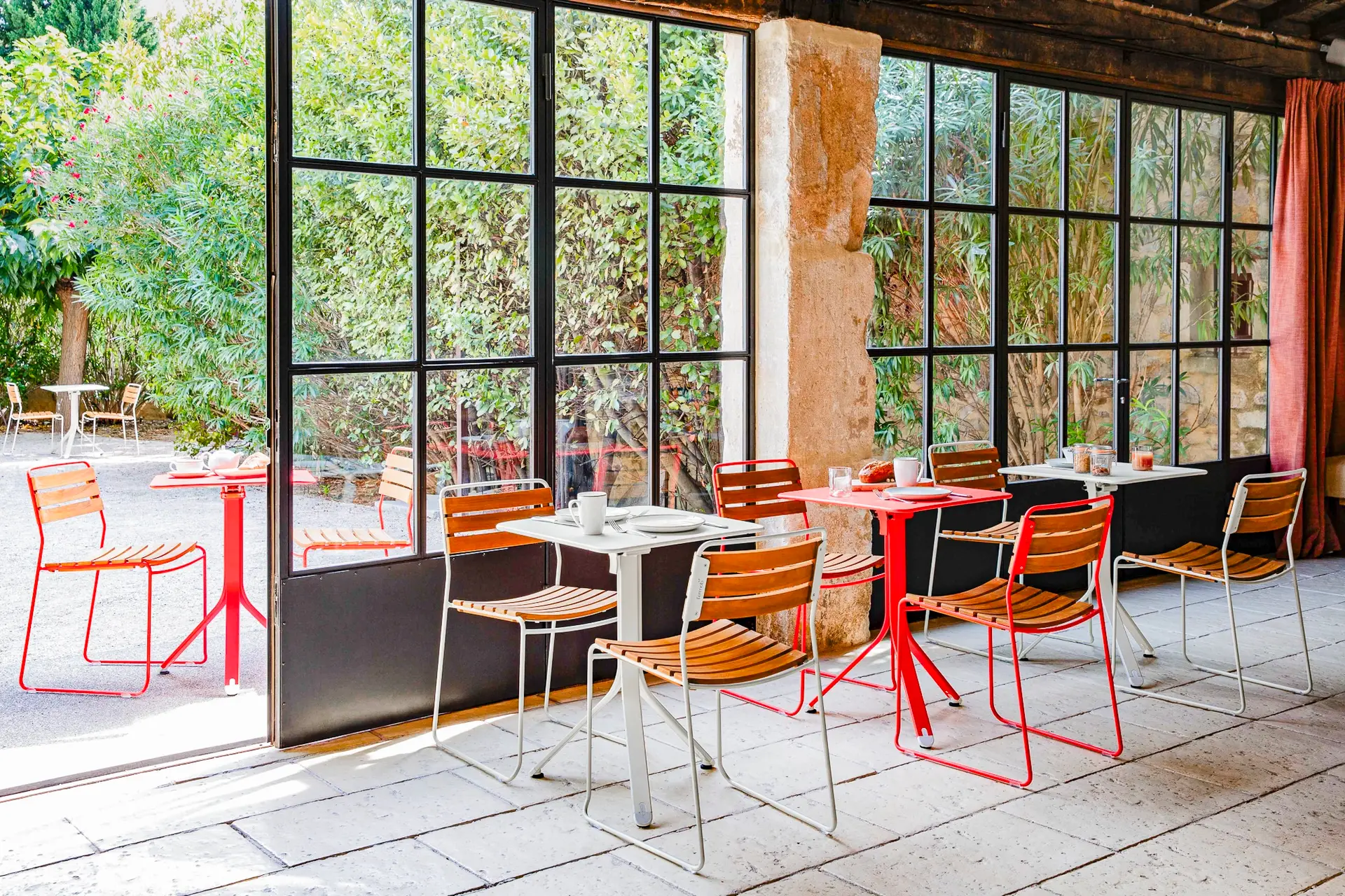 Ein lichtdurchflutetes Café mit roten und weißen Tischen, umgeben von Holzstühlen, bietet durch große Glasfenster einen Blick auf einen grünen Garten.