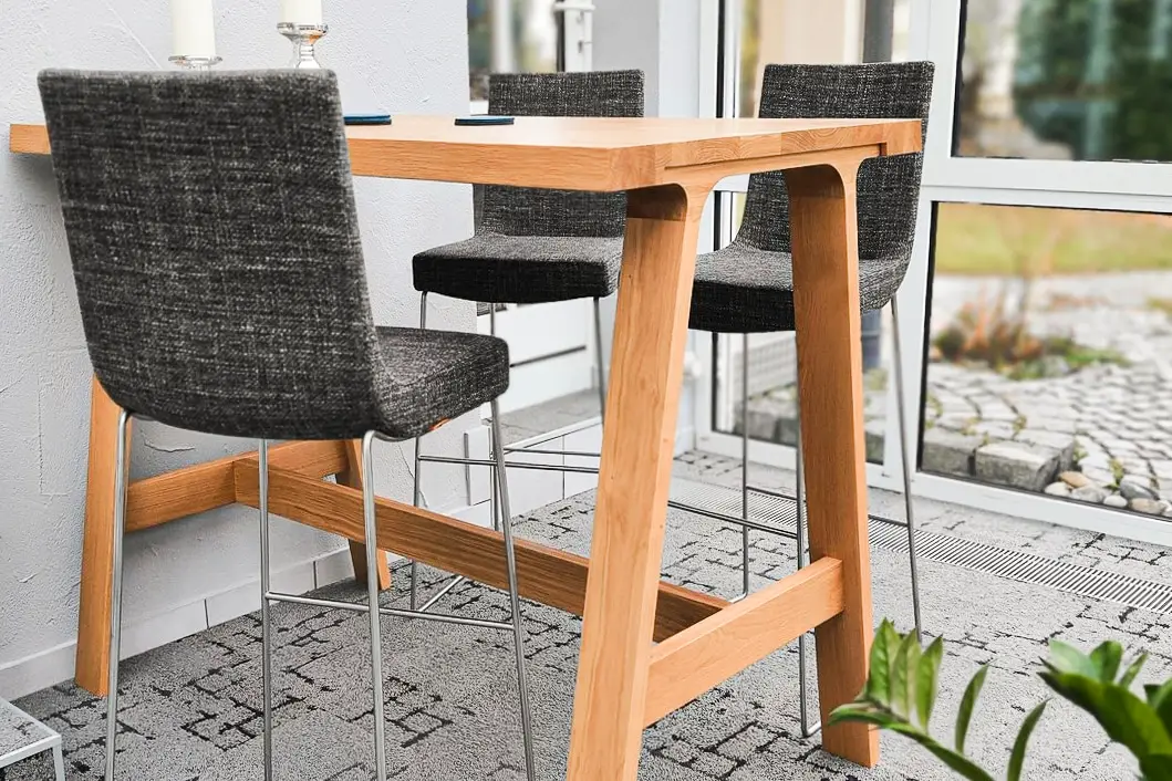 Eine Terrasse mit einem hohen Holztisch, drei grauen Stühlen und zwei weißen Kerzen auf silbernen Haltern. Neben der Tür stehen zwei weiße Laternen. Große Fenster lassen viel Licht herein.