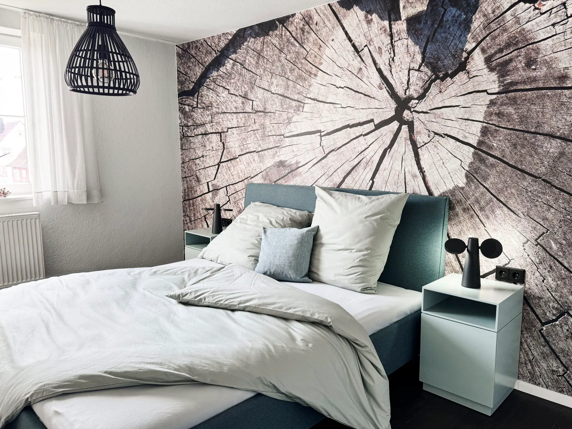 Referenz Nicole Hafen das wohnwerk, Schlafzimmer mit grauem Bett und Nachtischen, im Hintergrund befindet sich eine Tapete in Holzoptik