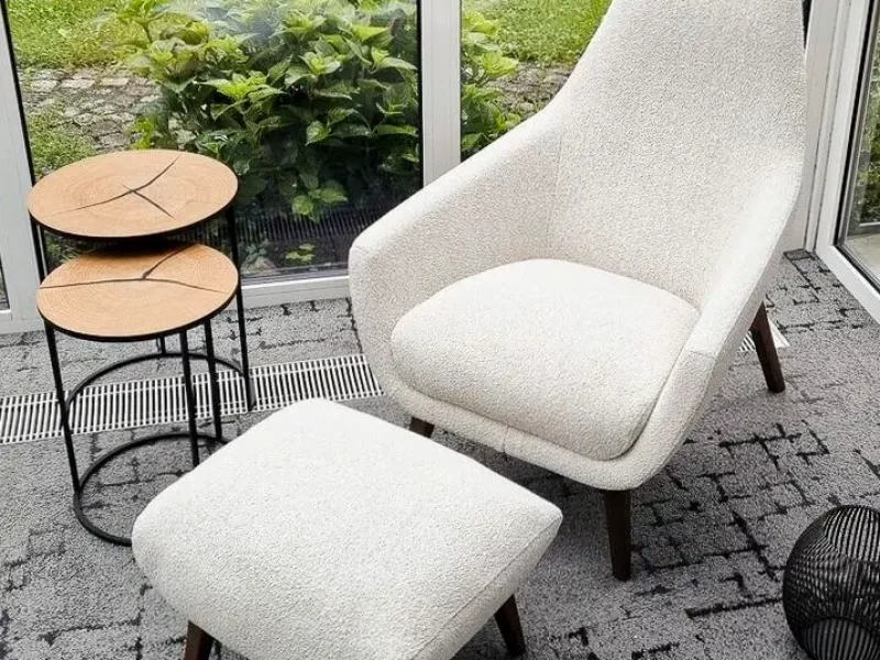 Ein heller Sitzbereich mit einem weißen Sessel, Fußhocker, zwei kleinen Holztischen und drei dekorativen Körben. Große Fenster bieten Blick auf den Garten.