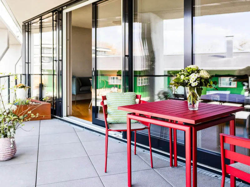 Balkon mit roten Gartenmöbeln. Links sind Pflanzen in Töpfen, und im Hintergrund ist eine große Glas-Schiebetür zu sehen, die den Balkon mit dem Innenraum verbindet.