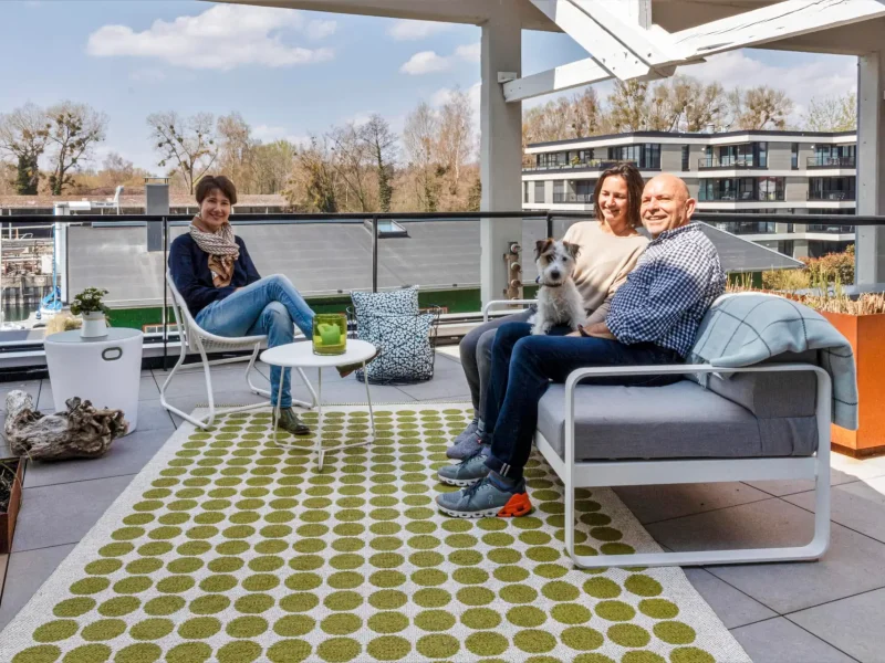 Frau Hafen und ein Pärchen mit einem Hund auf dem Schoß sitzen auf einem Balkon mit viel Dekoration. Sie sitzen auf modernen Möbeln, die auf einem weißen Teppich mit grünen Punkten stehen.