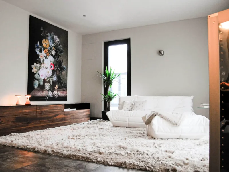 Ein modernes Wohnzimmer mit einem weißen Sofa, einem großen flauschigen Teppich und einer dunklen Holzkommode. An der Wand hängt ein großes Blumenbild. Eine grüne Pflanze steht neben einem Fenster, das Licht in den Raum bringt.
