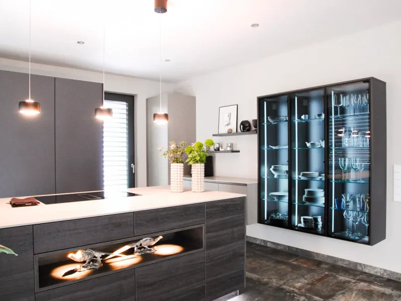 Eine moderne Küche mit grauen Schränken, einer Kücheninsel, drei schwarzen Pendelleuchten und einem beleuchteten Glasschrank für Geschirr und Gläser.