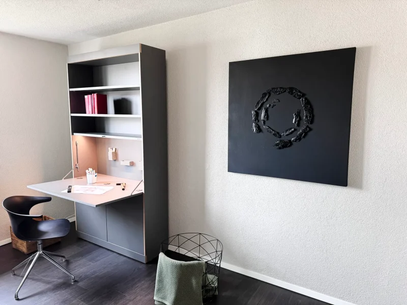 Ein modernes Arbeitszimmer mit einem schwarzen Stuhl an einem grauem Klapptisch im Regal. An der Wand hängt ein schwarzes Gemälde. Darunter steht ein Korb.