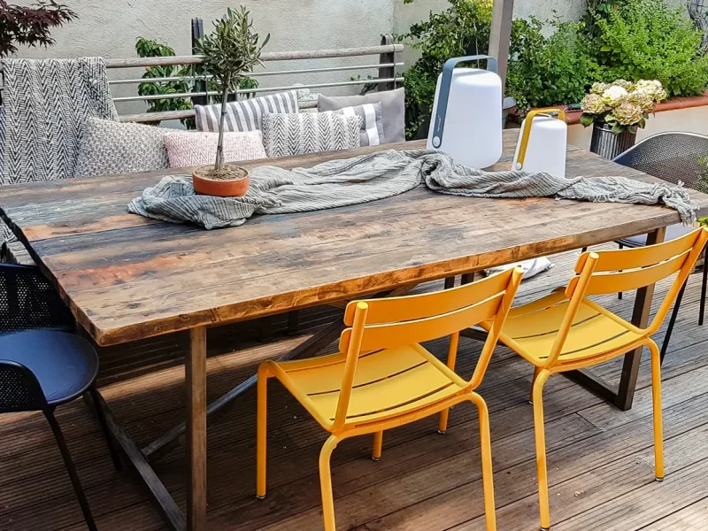 Eine gemütliche Terrasse mit einem rustikalen Holztisch, gelben und schwarzen Stühlen und zahlreichen Pflanzen. Auf dem Tisch liegen Decken und Lampen.