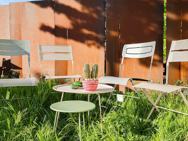 Eine Outdoor Sitzgruppe mit vier Stühlen und zwei kleinen runden Tischen mit Kakteendeko und weiß-rot gestreifter Vase im hohen grünen Gras