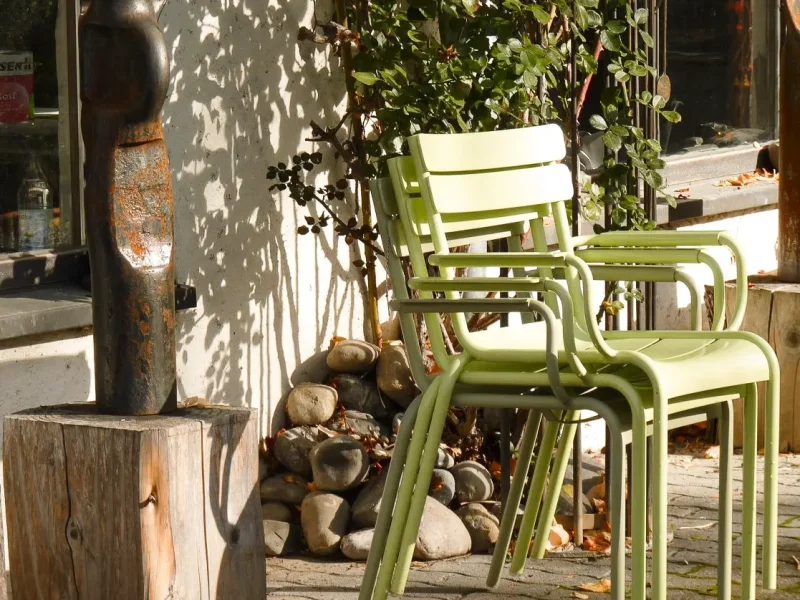 Gestapelte grüne Stühle stehen im Garten. Im Hintergrund sind Pflanzen, Steine und die Hauswand zu sehen.