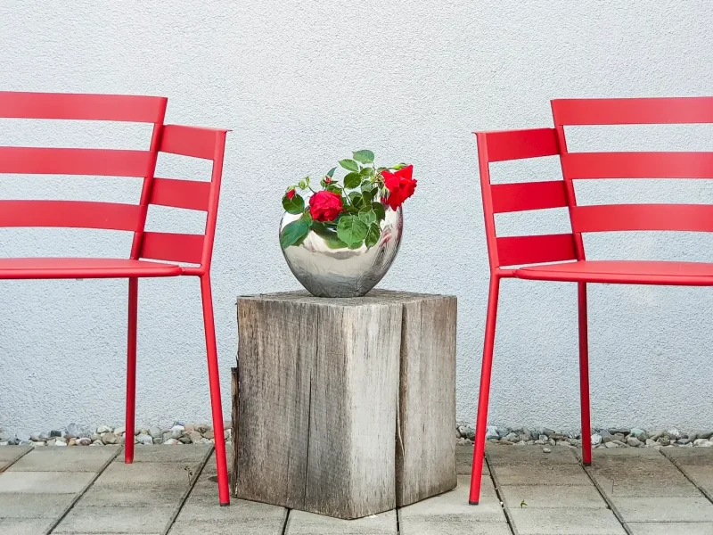 Zwei rote Metallstühle, die vor einer hellen Wand stehen. Zwischen den Stühlen befindet sich ein hölzerner Baumstumpf mit einem Blumentopf, der rote Blumen enthält.